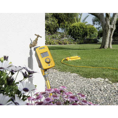 Ein gelber Gartenbewässerungstimer ist an einen Wasserhahn angeschlossen, während im Hintergrund ein Rasensprenger in einem Garten läuft.